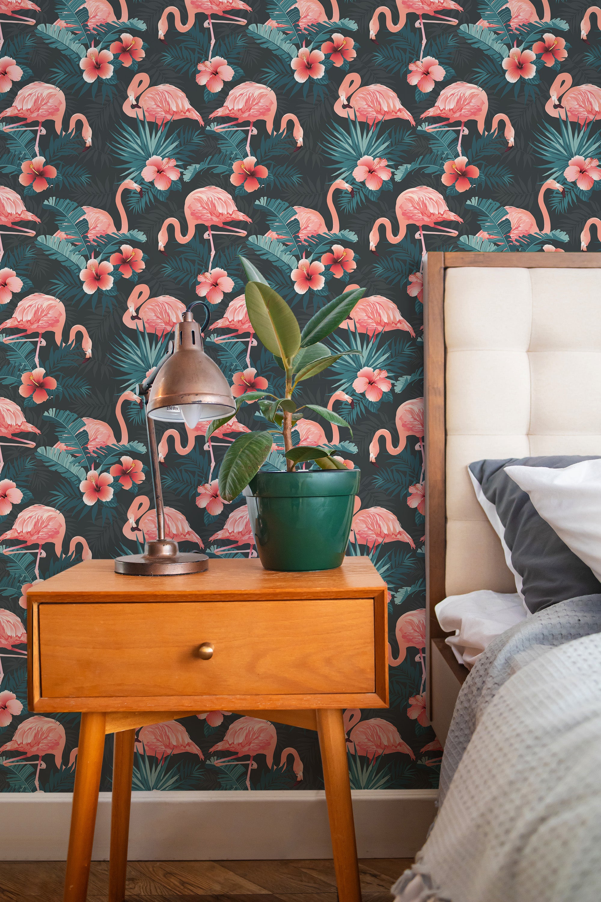 Self-adhesive wallpaper with vibrant pink flamingos and tropical leaves on a dark background, accented by pink flowers, installed behind a wooden bedside table with a lamp and potted plant.