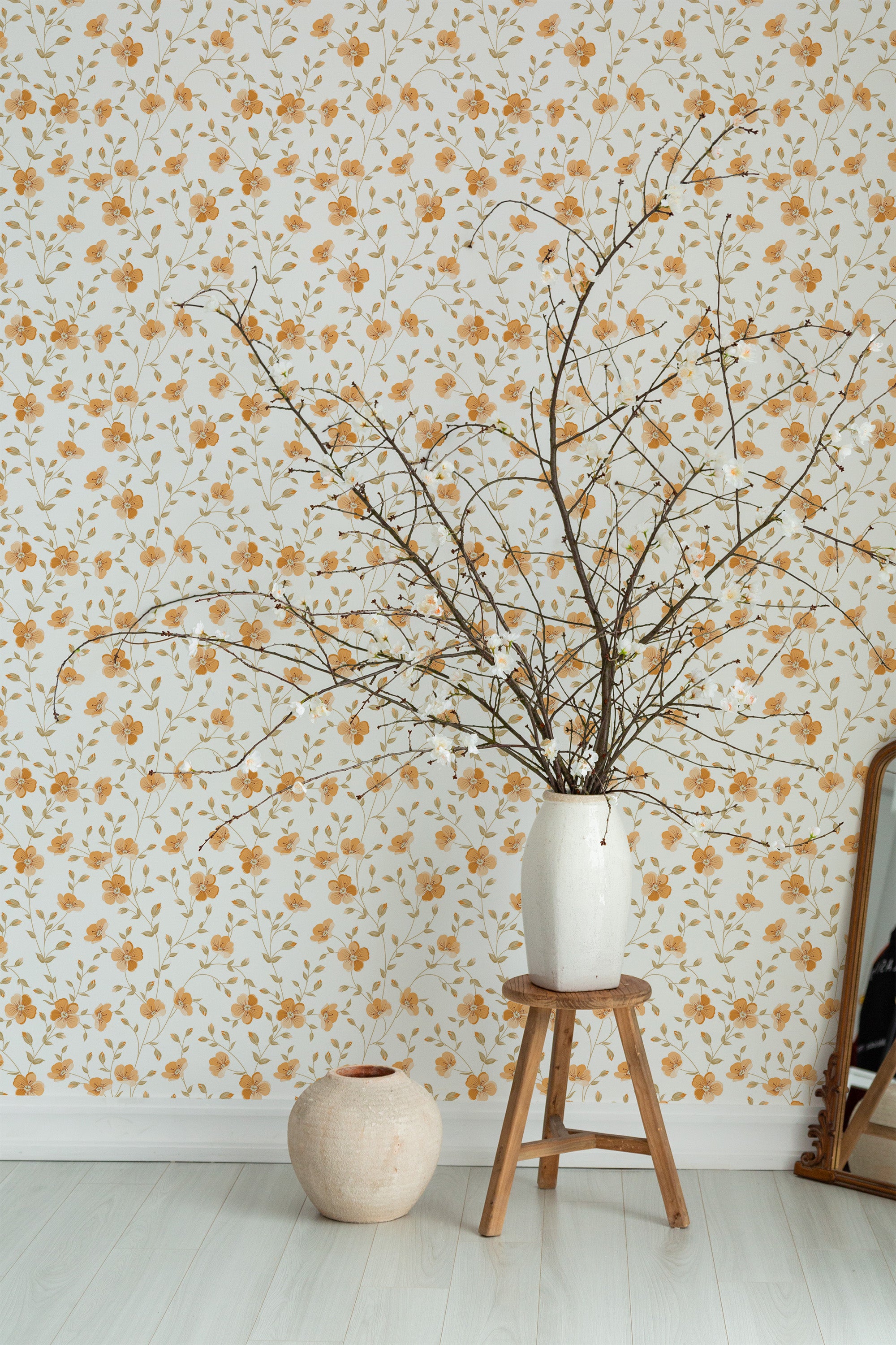 Self-adhesive wallpaper with an orange floral pattern on a light background adorning a room's wall, accompanied by a vase with branches and a small stool.