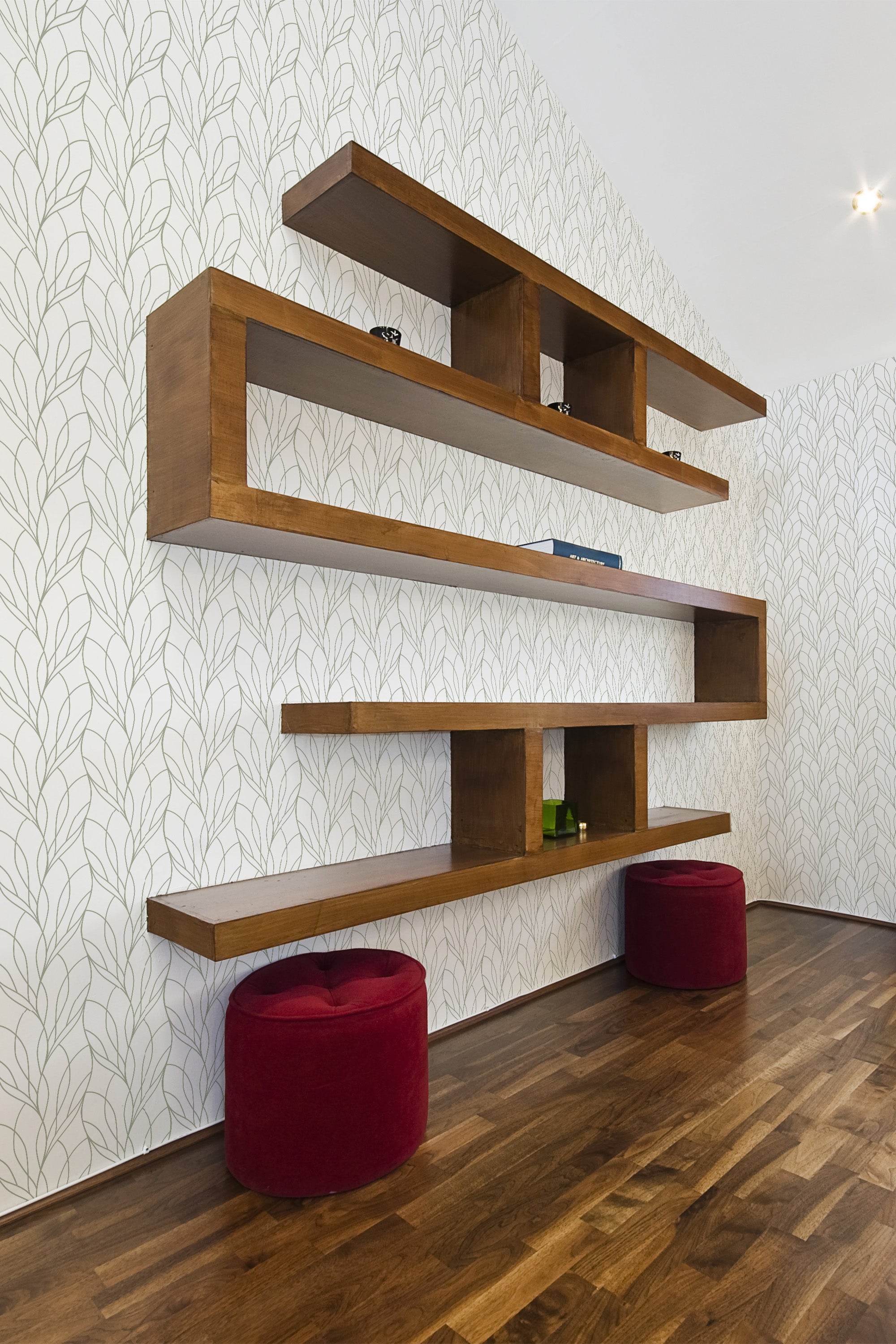 A room with a wall covered in wavy line patterned wallpaper in neutral tones, complemented by modern wooden shelves and two red ottomans, reflecting a blend of contemporary and traditional wallpaper styles.