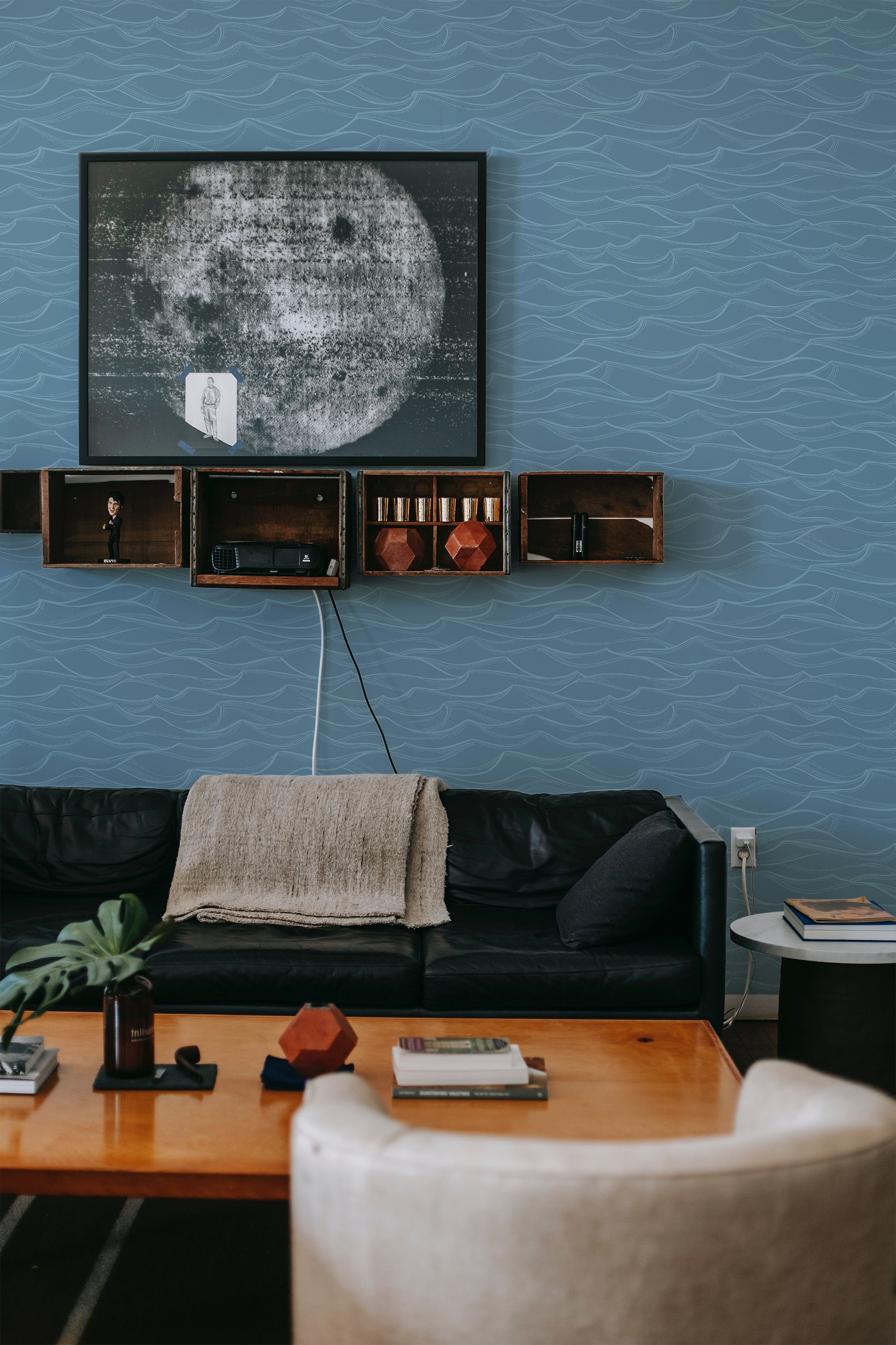 Blue wallpaper with subtle, serene wave patterns mimicking the gentle movement of the ocean, bringing a calm, airy atmosphere to the room. The design complements the dark leather sofa and minimalist wooden shelves, adding a touch of nature-inspired elegance to this space. Traditional wallpaper.