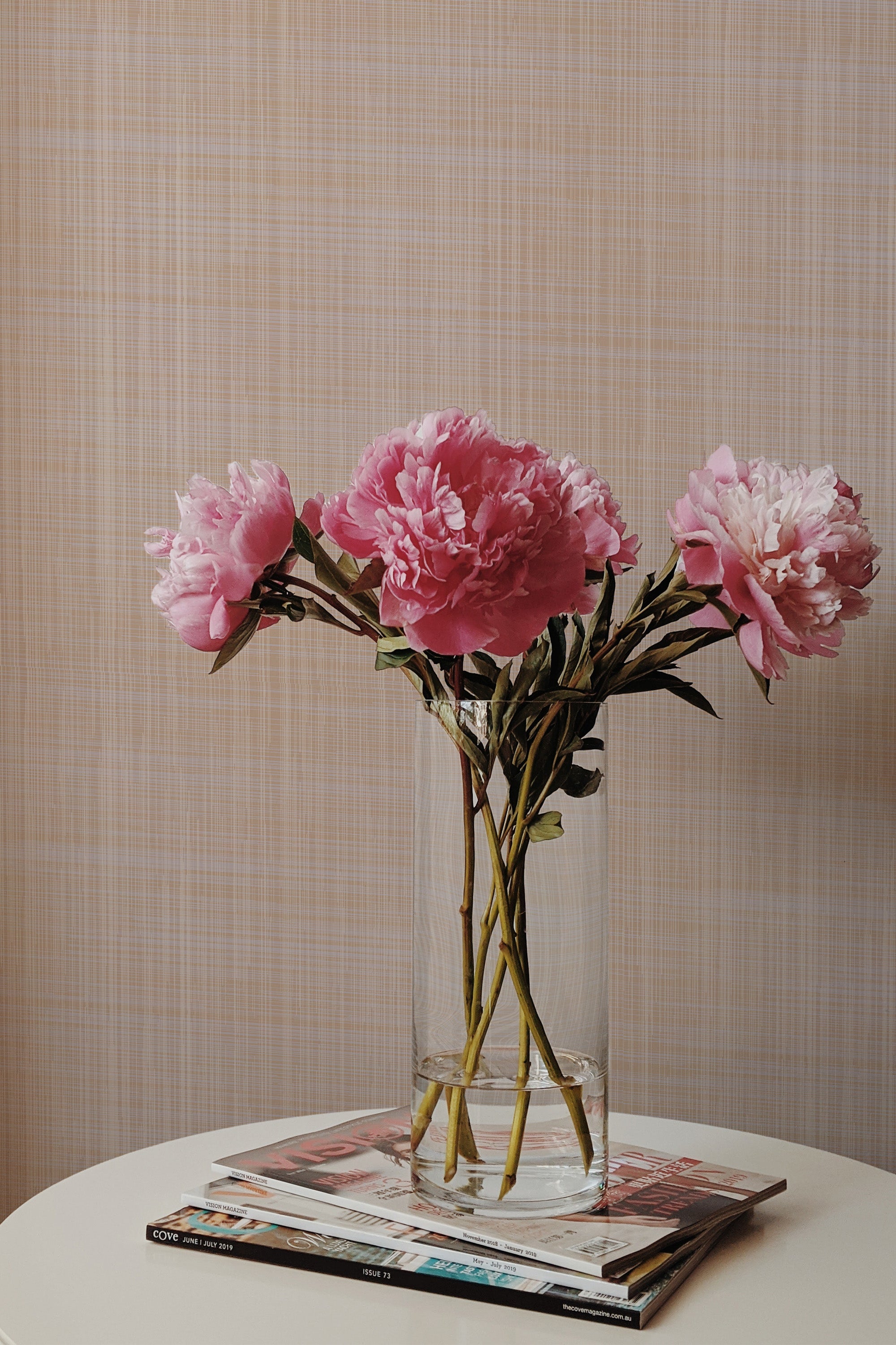 Self-adhesive faux tweed fabric wallpaper with subtle herringbone pattern in a room, accented by a vase of pink peonies on a table.
