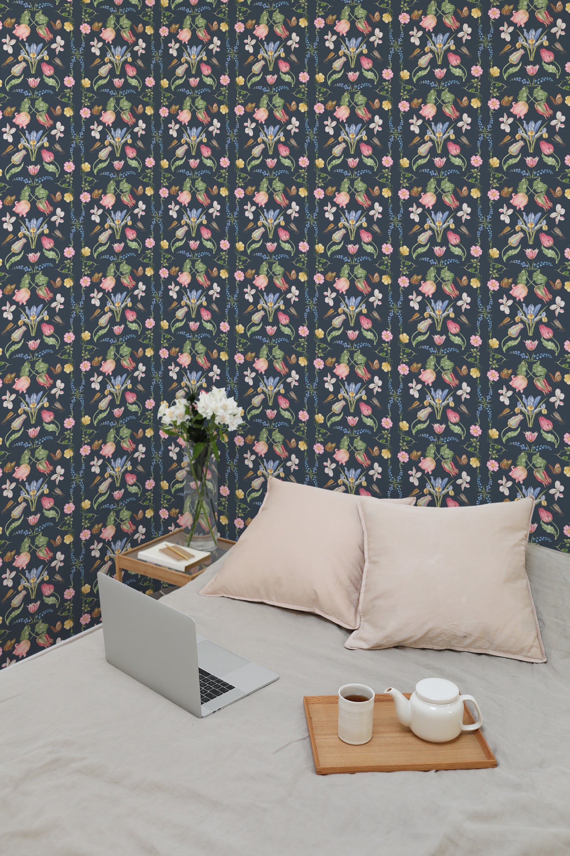Self-adhesive dark ornamented floral wallpaper with pink and blue flowers on a navy background, in a room with a laptop and cushions on the bed.