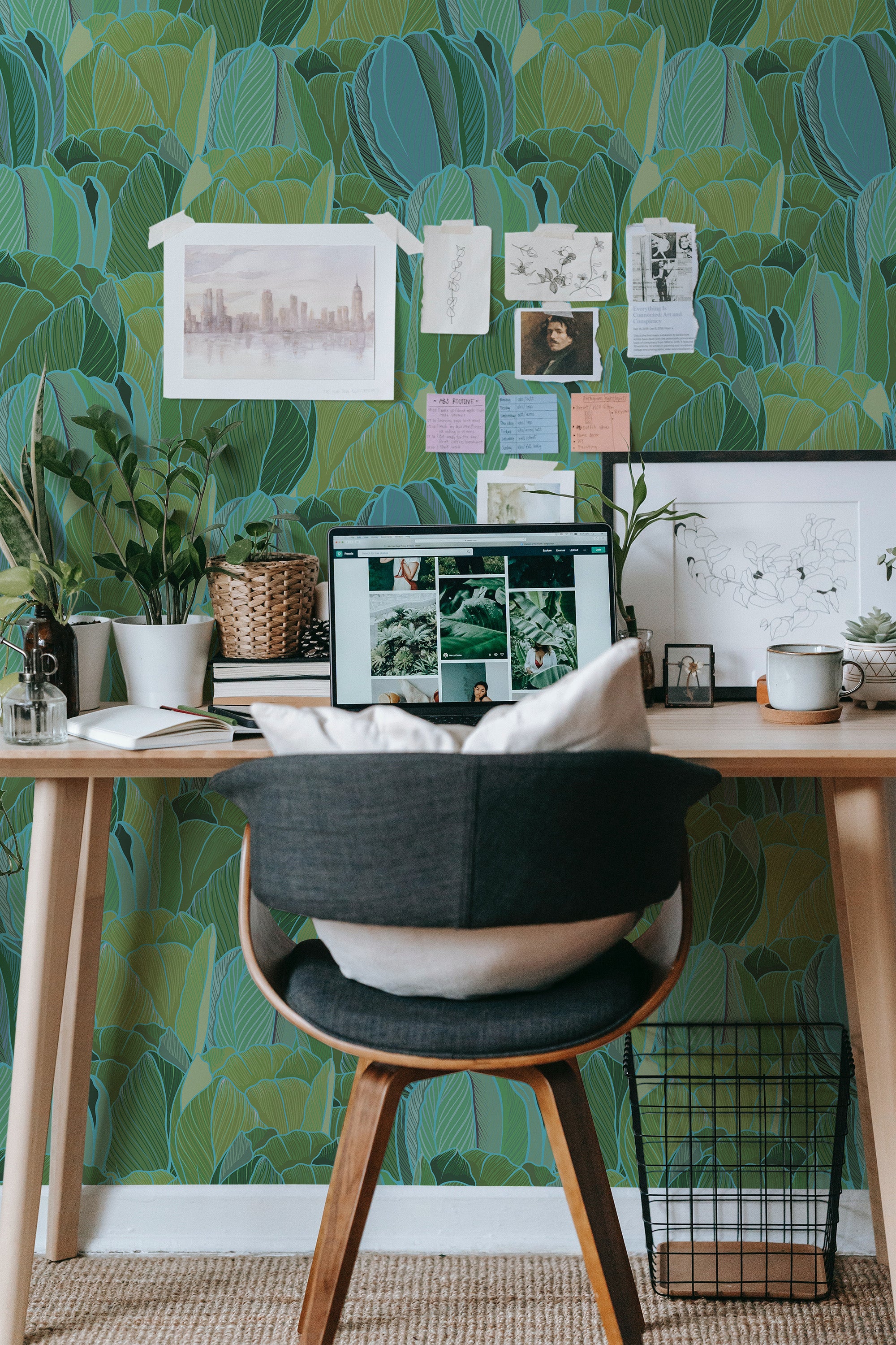 Self-adhesive wallpaper with a pattern of green and teal leaves in a room with a wooden desk, a chair, potted plants, and various papers tacked to the wall.