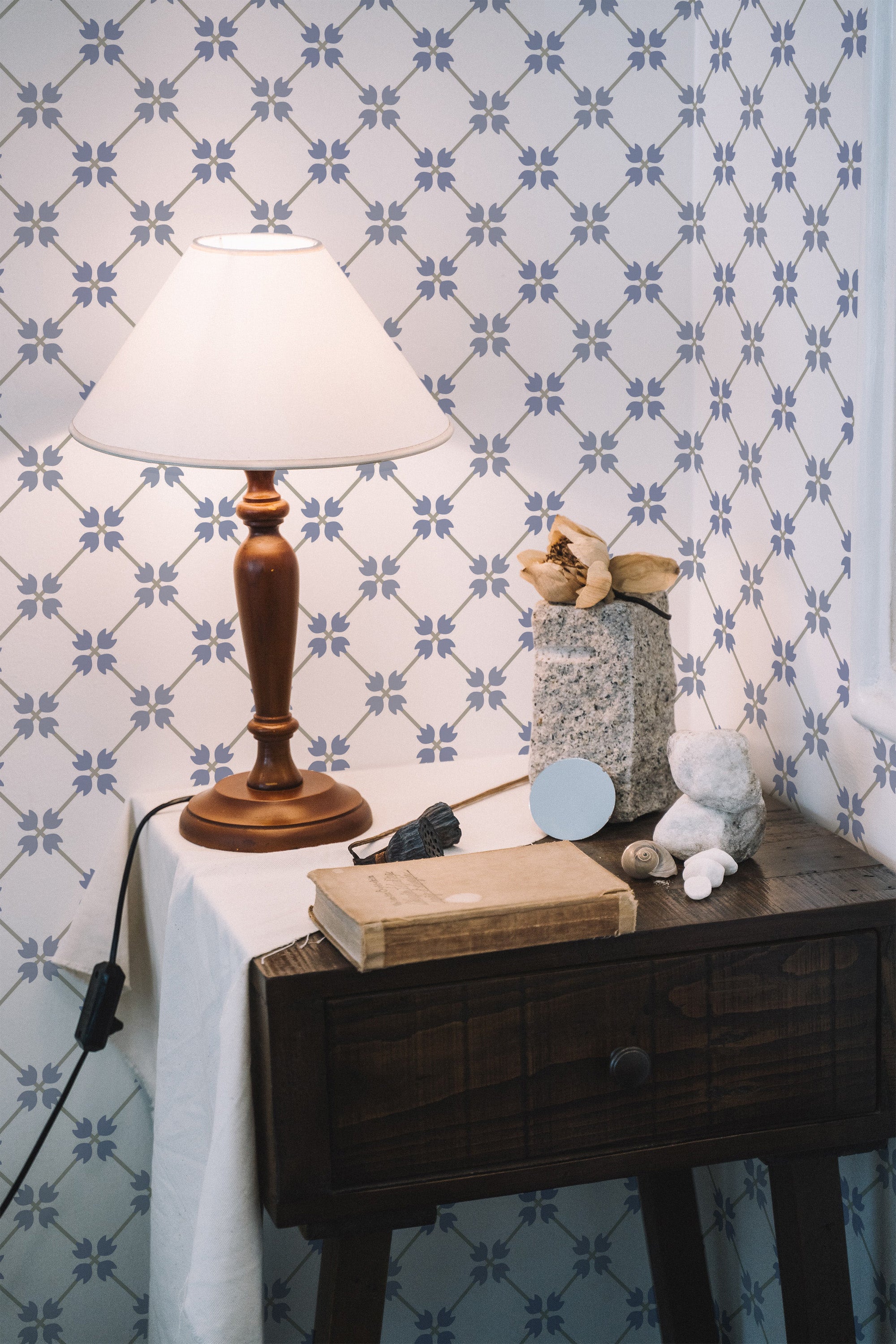 Self-adhesive wallpaper with a retro grid pattern composed of small, stylized blue floral motifs arranged in a symmetrical diamond layout, creating a vintage and elegant look. The subtle backdrop complements the wooden side table, lamp, and decorative items in the room.