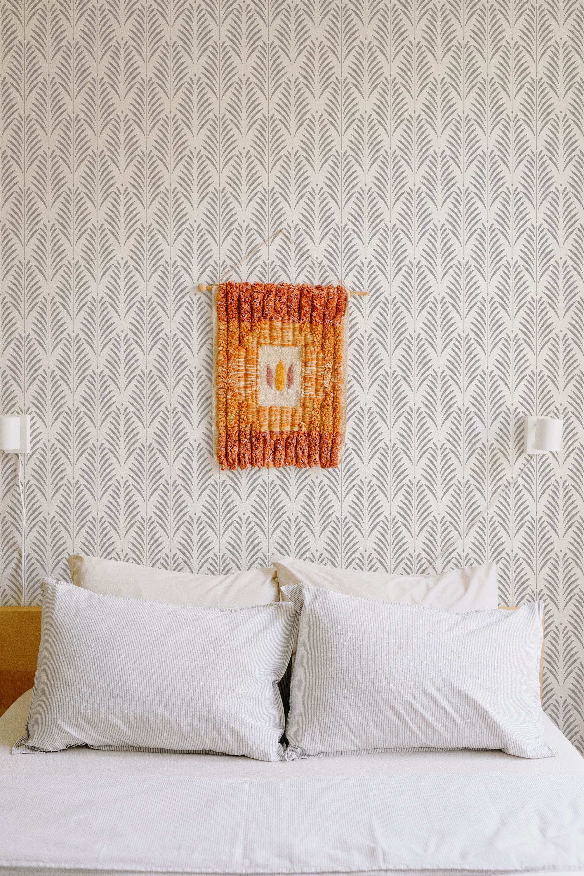 Self-adhesive wallpaper with a repetitive leaf pattern in a neutral color scheme on a bedroom wall, above a bed with white pillows.
