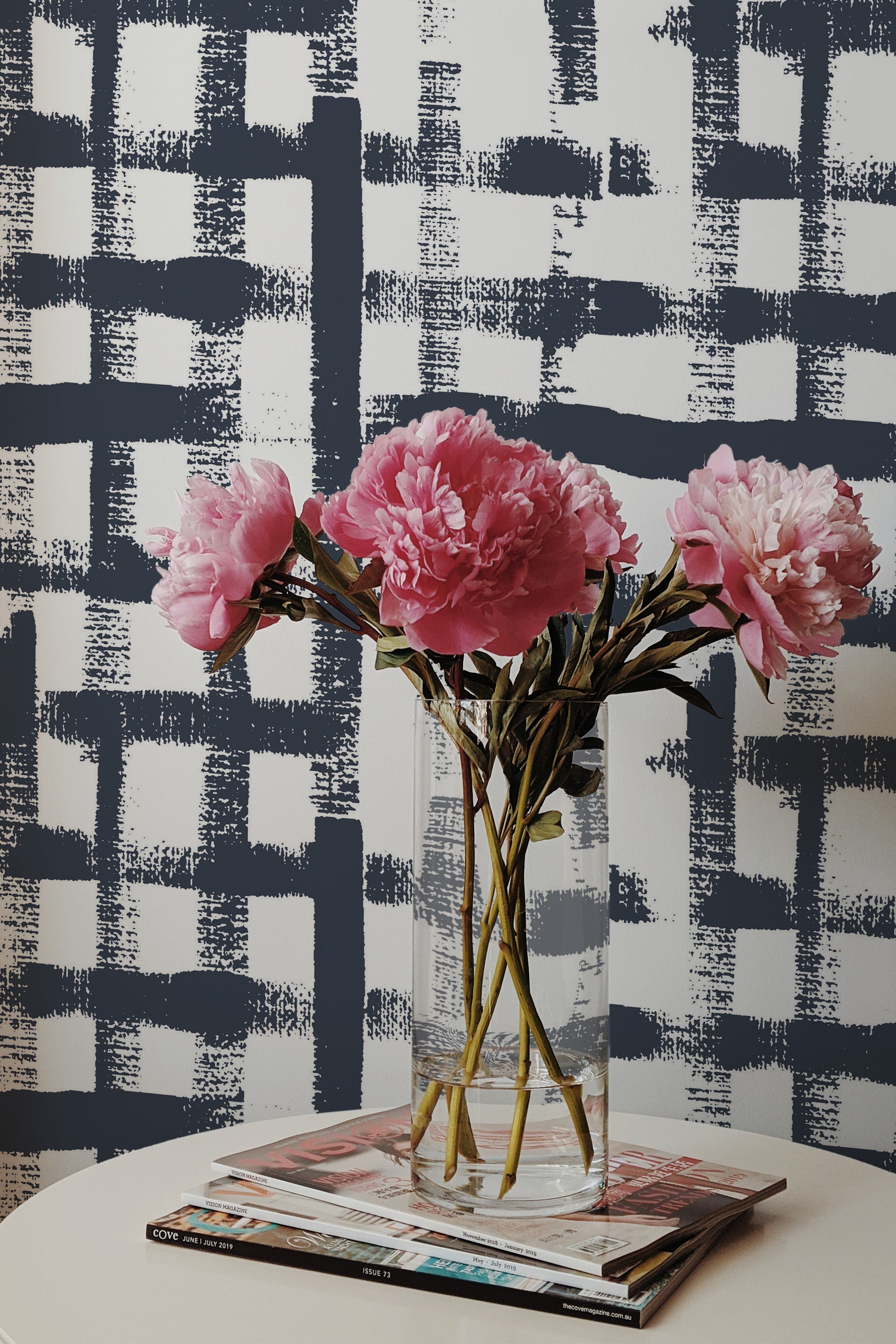 Self-adhesive wallpaper with a Navy blue brush stroke design on a light background, creating a modern and dynamic grid pattern. Pink flowers in a clear vase are placed in front of the wallpaper, adding a touch of softness to the room's d��cor.