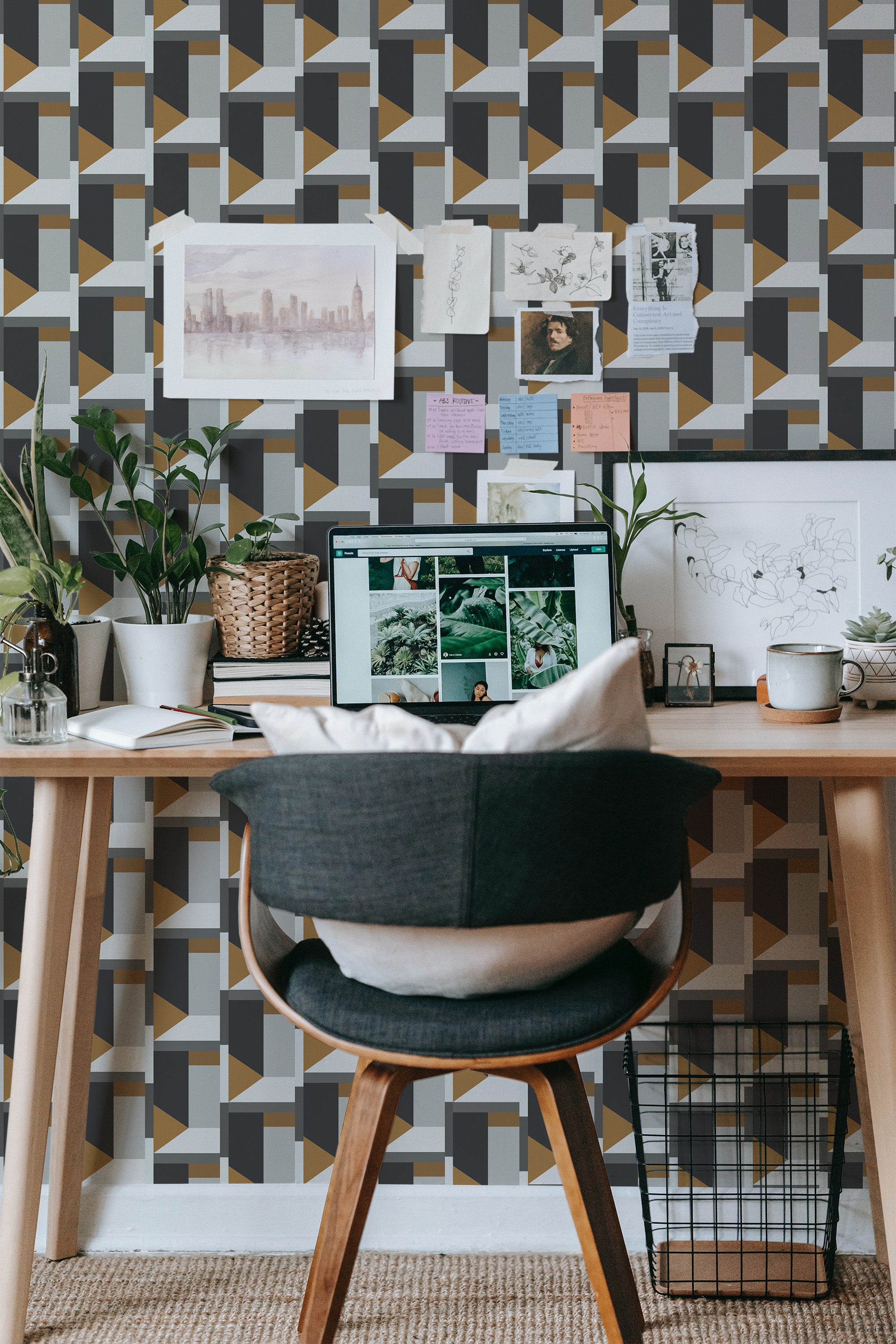 Self-adhesive luxury geometric wallpaper with an intricate pattern in shades of black, gray, white, and gold. The design includes interlocking shapes and creates a bold, modern appearance behind a cozy, well-organized workspace with plants, art, and notes on the wall.
