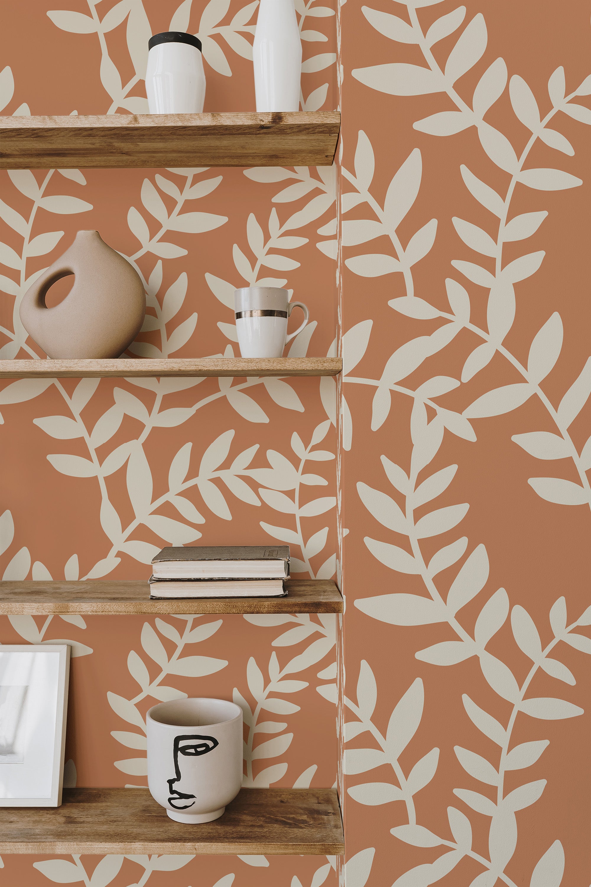 Self-adhesive wallpaper with a neutral leaf bush design on a warm terracotta backdrop, shown behind wooden shelves with various decorative items.